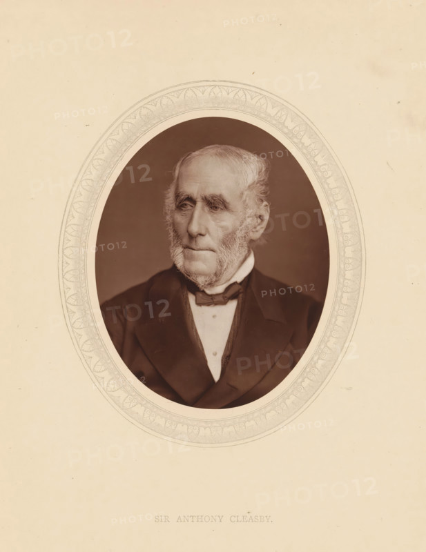 Sir Anthony Cleasby, page, photomechanical print, photographer: Lock & Whitfield, cliché maker: anonymous, c. 1875 - in or before 1880, paper, Woodburytype, Woodbury Process, height 115 mm x width 89 mm, Person, Anthony Claesby, magistracy, judges, historical persons (portraits and scenes from the life) ((full) bust portrait), An oval-framed sepia portrait of an elderly man with light hair and prominent sideburns, wearing a dark suit jacket, a white shirt, and a bow tie, set against a plain background; the oval frame has a decorative patterned border and the portrait is centered on a light-colored sheet with some small blemishes and spots.