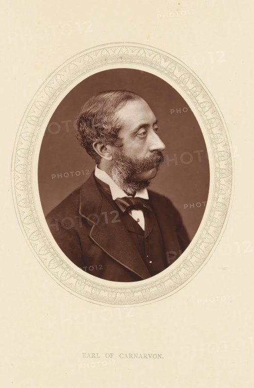 Earl of Carnarvon, page, photomechanical print, photographer: Lock & Whitfield, cliché maker: anonymous, c. 1875 - in or before 1880, paper, Woodburytype, Woodbury Process, height 115 mm x width 89 mm, Person, Henry Herbert, politician, eg party leader, historical persons (portraits and scenes from the life) ((full) bust portrait), A sepia oval portrait of a man in three-quarter profile facing right, framed by a decorative patterned border set on a light background, showing the man's wavy short hair, prominent mustache and sideburns, high forehead, and contemplative expression; he wears a dark jacket with a visible lapel, a white shirt collar, a bow tied at the neck, and a waistcoat beneath, all photographed against a plain dark backdrop within the ornate oval frame.