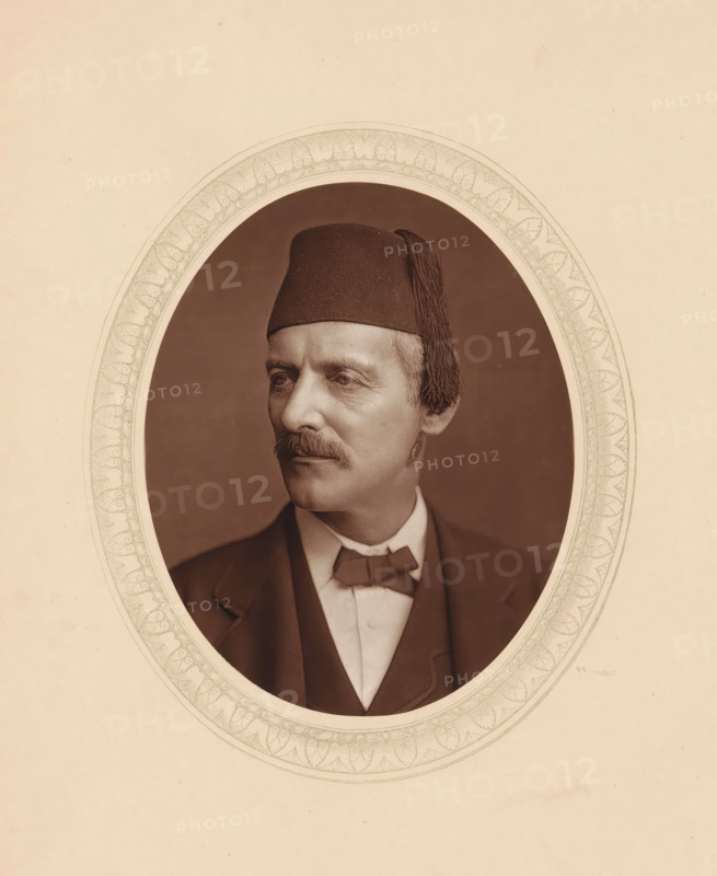 William Gifford Palgrave, page, photomechanical print, photographer: Lock & Whitfield, cliché maker: anonymous, c. 1875 - in or before 1880, paper, Woodburytype, Woodbury Process, height 115 mm x width 89 mm, Person, Gifford Palgrave, scholar, philosopher, historical persons (portraits and scenes from the life) ((full) bust portrait), A sepia-toned oval portrait of a middle-aged man wearing a dark tasseled fez and a suit with a bow tie, his face turned slightly to the side showing a trimmed mustache and short hair, set against a plain dark background and framed by an ornate decorative border on a light paper background.