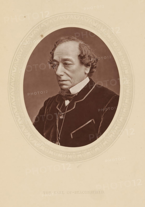 The earl of Beaconsfield, page, photomechanical print, photographer: Downey, William & Daniel, cliché maker: anonymous, c. 1876 - in or before 1881, paper, Woodburytype, Woodbury Process, height 115 mm x width 89 mm, Person, Benjamin Disraeli, politician, eg party leader, historical persons (portraits and scenes from the life) ((full) bust portrait), A sepia-toned oval portrait of an older man with wavy hair and sideburns, shown from the chest up and turned slightly to his left, wearing a dark coat with light piping, a high white collar, a bow tie, and a pendant on a chain; the portrait is set within a decorative oval frame on a pale background.