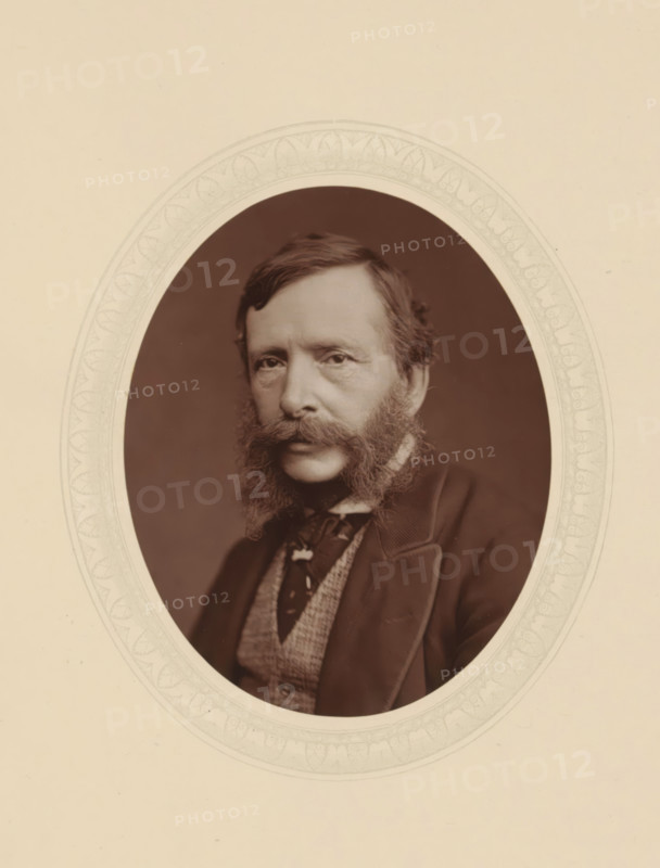 William Charles T. Dobson, page, photomechanical print, photographer: Lock & Whitfield, cliché maker: anonymous, c. 1876 - in or before 1881, paper, Woodburytype, Woodbury Process, height 115 mm x width 89 mm, Person, William Charles Thomas Dobson, portrait, self-portrait of painter, historical persons (portraits and scenes from the life) ((full) bust portrait), A sepia portrait of a man shown within an ornate oval frame against a light beige background, the man has a full styled beard and prominent sideburns, short neatly combed hair, and a solemn facial expression; he wears a dark jacket with a visible lapel, a patterned waistcoat, and a necktie with a decorative knot, and the oval mat around the portrait has a faint decorative leaf-and-scroll border.