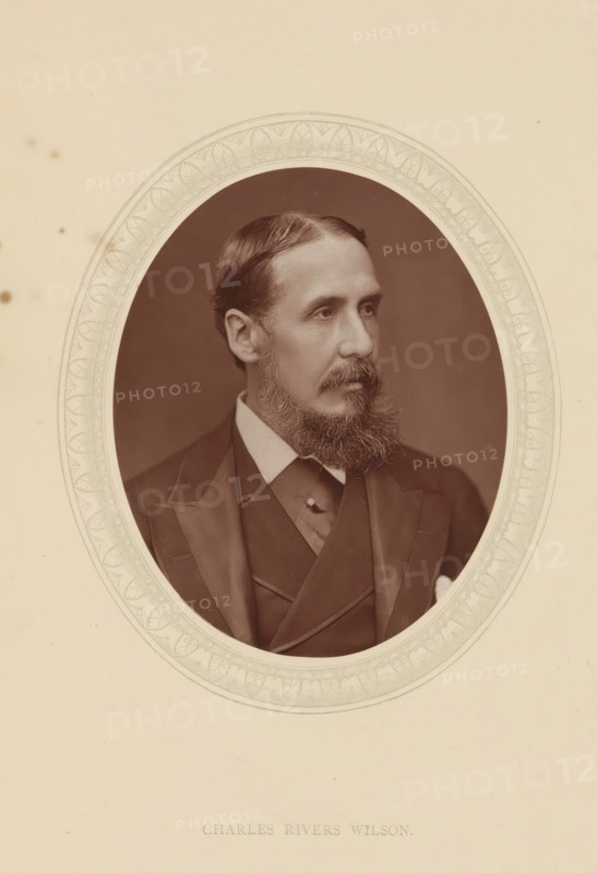 Charles Rivers Wilson, page, photomechanical print, photographer: Lock & Whitfield, cliché maker: anonymous, c. 1876 - in or before 1881, paper, Woodburytype, Woodbury Process, height 115 mm x width 89 mm, Person, official, civil servant, historical persons (portraits and scenes from the life) ((full) bust portrait), A sepia-toned oval portrait of a man with neatly combed hair and a full beard, shown from the chest up and turned slightly to the side, wearing a dark suit with a high-collared shirt and a waistcoat, set against a plain dark background and surrounded by an ornate patterned oval border on a light-colored mat.