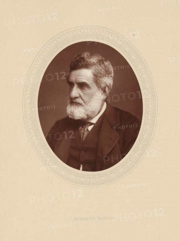Hormuzd Rassam, FRGS, page, photomechanical print, photographer: Lock & Whitfield, cliché maker: anonymous, c. 1876 - in or before 1881, paper, Woodburytype, Woodbury Process, height 115 mm x width 89 mm, Person, diplomacy, diplomat, historical persons (portraits and scenes from the life) ((full) bust portrait), An oval sepia portrait of an older man with a full white beard and moustache, short wavy hair, and a serious expression, shown in three-quarter profile facing left, wearing a dark suit jacket, waistcoat, collared shirt, and tie, set against a plain dark background and framed within a decorative oval mat on a light beige page.