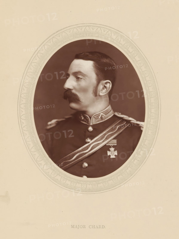 Major Chard, page, photomechanical print, photographer: Lock & Whitfield, cliché maker: anonymous, c. 1876 - in or before 1881, paper, Woodburytype, Woodbury Process, height 115 mm x width 89 mm, Person, John Chard, officer, historical persons (portraits and scenes from the life) ((full) bust portrait), A sepia oval portrait of a man in a military uniform shown in profile facing left, with dark hair combed back and a thick mustache, wearing a high-collared jacket with decorative trim and metal buttons, a sash across his chest with braided cords on the shoulder, and a medal pinned to the jacket, all set against a plain darker background and framed by an ornate patterned oval mat on a light-colored page.