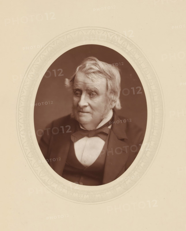 Right hon. JA Roebuck, page, photomechanical print, photographer: Lock & Whitfield, cliché maker: anonymous, c. 1876 - in or before 1879-11, paper, Woodburytype, Woodbury Process, height 115 mm x width 89 mm, Person, John Arthur Roebuck, politician, eg party leader, historical persons (portraits and scenes from the life) ((full) bust portrait), An oval sepia portrait of an older person with light hair and sideburns set against a dark background, framed by a decorative pale border on a light beige matte; the subject wears a dark coat over a vest and a bow at the collar, and their expression is neutral with eyes looking slightly downward and to the side.