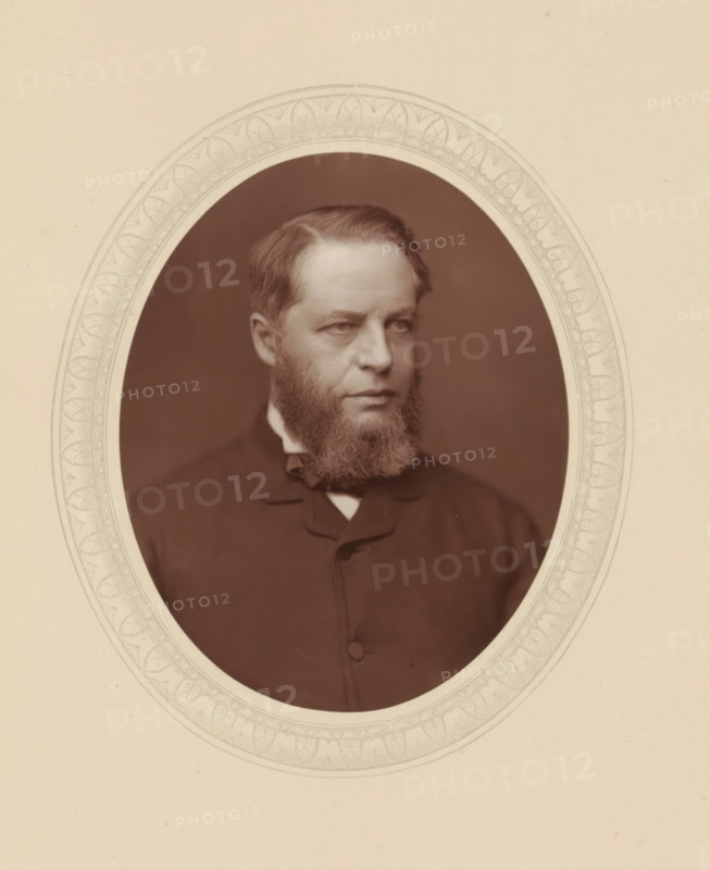 Sir Henry Charles Lopes, page, photomechanical print, photographer: Lock & Whitfield, cliché maker: anonymous, c. 1876 - in or before 1881, paper, Woodburytype, Woodbury Process, height 115 mm x width 89 mm, Person, Henry Lopes, magistracy, judges, historical persons (portraits and scenes from the life) ((full) bust portrait), A sepia-toned oval portrait of a middle-aged man with a full beard and short hair, shown from the chest up and turned slightly to his left, wearing a dark coat with a high collar and a visible bow at the neck, set against a dark background and framed by a decorative oval mat with a patterned inner border on a light background.
