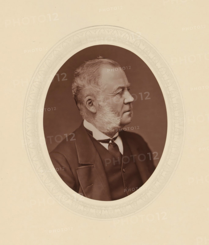 Duke of Richmond and Gordon, page, photomechanical print, photographer: Lock & Whitfield, cliché maker: anonymous, c. 1877 - in or before 1882, paper, Woodburytype, Woodbury Process, height 115 mm x width 89 mm, Person, Charles Gordon-Lennox, politician, eg party leader, historical persons (portraits and scenes from the life) ((full) bust portrait), A sepia-toned oval portrait of an older man shown in profile facing right, with short swept-back hair and prominent sideburns, wearing a dark suit jacket, waistcoat, collared shirt, and a tied neckwear; the portrait is centered within a decorative oval mat with a patterned border and set against a plain light background.