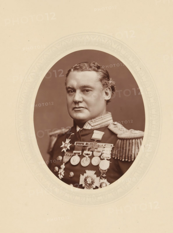Sir William Hewett, page, photomechanical print, photographer: Lock & Whitfield, cliché maker: anonymous, c. 1877 - in or before 1882, paper, Woodburytype, Woodbury Process, height 115 mm x width 89 mm, Person, William Nathan Wrighte Hewett, officer, historical persons (portraits and scenes from the life) ((full) bust portrait), An oval sepia portrait shows a man from the chest up wearing a dark military uniform with ornate shoulder epaulettes and a high collar; he has wavy hair combed back and a composed facial expression, and his chest is adorned with multiple medals and decorative insignia; the portrait is set against a plain darker background and framed by a patterned oval mat on a light-colored mount.