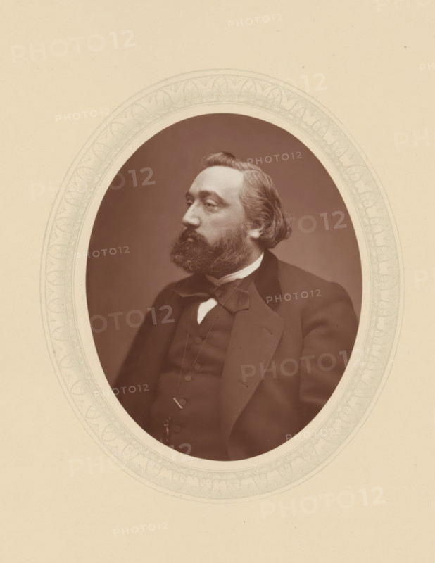 Léon Gambetta, page, photomechanical print, photographer: Lock & Whitfield, cliché maker: anonymous, c. 1877 - in or before 1882, paper, Woodburytype, Woodbury Process, height 115 mm x width 89 mm, Person, prime minister, historical persons (portraits and scenes from the life) ((full) bust portrait), An oval sepia photograph of a bearded man shown from the chest up, wearing a dark suit with a bow tie and waistcoat, his hair swept back and a full beard, photographed in profile facing left, set inside a decorative oval mat with a pale patterned border against a plain light background.