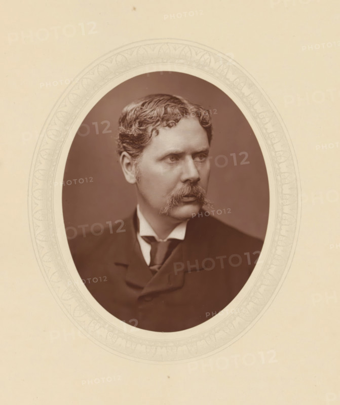 Marcus Stone, page, photomechanical print, photographer: Lock & Whitfield, cliché maker: anonymous, c. 1877 - in or before 1882, paper, Woodburytype, Woodbury Process, height 115 mm x width 89 mm, Person, portrait, self-portrait of painter, historical persons (portraits and scenes from the life) ((full) bust portrait), An oval sepia photograph of a man with wavy hair and a prominent mustache is centered on a pale background, the man shown from the chest up wearing a dark jacket, high collared shirt, and tie, his gaze directed slightly to the side, and the oval photo is bordered by a faint decorative pattern on the surrounding mat.