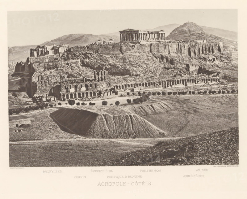 View of the Acropolis of Athens, Acropole-côté S, page, photomechanical print, inscription: 'Propylées, Asklépiéion', Athens, cliché maker: Dujardin, printer (person): Chardon-Wittmann, c. 1880 - in or before 1890, photographer, printer (person), Periféreia Protevoúsis, paper, photoaquatint, height 146 mm x width 213 mm, Print on frontispiece., city-view in general, 'veduta', Akropolis, A wide sepia-toned photograph of an elevated rocky plateau topped by ancient stone ruins, showing rows of tall columns and rectangular structures along the plateau edge, with steep rocky slopes descending to a lower terrace that contains a long series of arched stone openings and scattered ruined walls; in front of the terrace there are small trees aligned along a pathway and cultivated plots with furrowed earth and low ridges, while the foreground shows open textured fields and a deep cutaway of earth; distant rounded hills form the background under a pale sky.