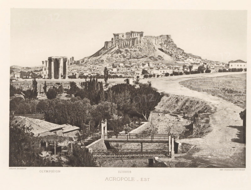 View of the Acropolis of Athens, Acropole-est, page, photomechanical print, Athens, cliché maker: Dujardin, printer (person): Chardon-Wittmann, c. 1880 - in or before 1890, photographer, printer (person), Periféreia Protevoúsis, paper, photoaquatint, height 145 mm x width 215 mm, city-view in general, 'veduta', ruin of a building, architecture, Akropolis, A wide sepia-toned landscape showing a rocky hill topped by ancient columned ruins overlooking a town with clustered low buildings and scattered trees, a group of tall standing columns visible on the plain near the hill, a winding dry riverbed running through the scene, foreground vegetation and tiled rooftops, and a pair of iron footbridges crossing a sunken channel.