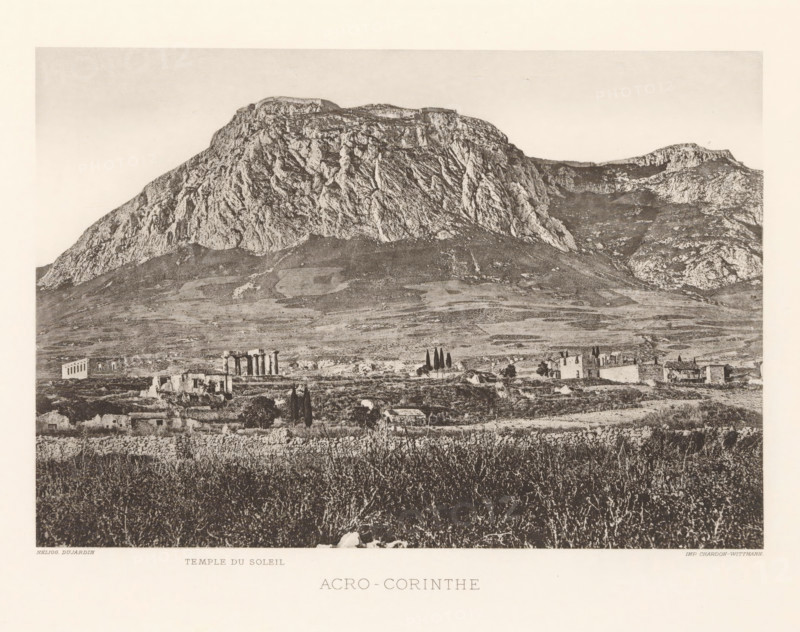Acro-Corinth, View of the Acrokorinthos, page, photomechanical print, inscription: 'Temple du soleil', Pelopónnesos, cliché maker: Dujardin, printer (person): Chardon-Wittmann, c. 1880 - in or before 1890, photographer, printer (person), paper, photoaquatint, height 146 mm x width 213 mm, landscape with ruins, mountains, Akrokorinthos, A large rocky mountain dominates the upper portion of the image with steep textured cliffs and a flatter ridge along its top, lighter sky above, and a gently sloping plains area below showing patches of vegetation and rocky ground; scattered in the midground are small stone ruins and buildings with vertical column remnants and taller slender trees nearby, while the foreground shows dense grasses and low shrubs with a rough stone wall along the lower edge.