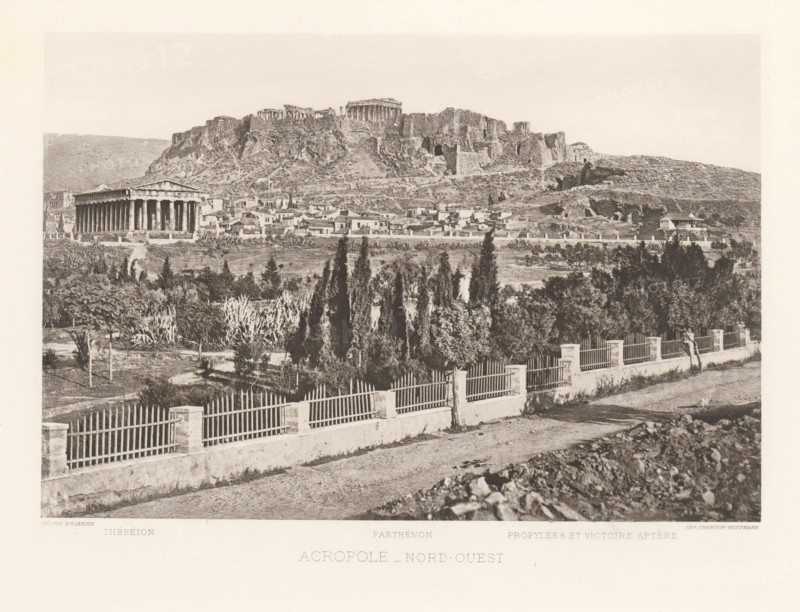 View of the Acropolis of Athens, Acropole- nord-ouest, page, photomechanical print, Athens, cliché maker: Dujardin, printer (person): Chardon-Wittmann, c. 1880 - in or before 1890, photographer, printer (person), Periféreia Protevoúsis, paper, photoaquatint, height 145 mm x width 215 mm, city-view in general, 'veduta', Akropolis, A wide sepia-toned view shows a rocky hill topped by ancient ruins with columns and stone structures, a lower area of clustered small buildings and houses at the hill's base, and a prominent rectangular temple with a colonnade standing apart from the houses. In the foreground a road runs parallel to a low wall topped by evenly spaced vertical railings and short square posts, behind which a planted area contains tall slender trees, dense shrubs, and other vegetation. The middle distance contains open grassy patches, scattered trees, and terraces leading up to the rugged slope of the hill, while the sky above is pale and mostly empty.