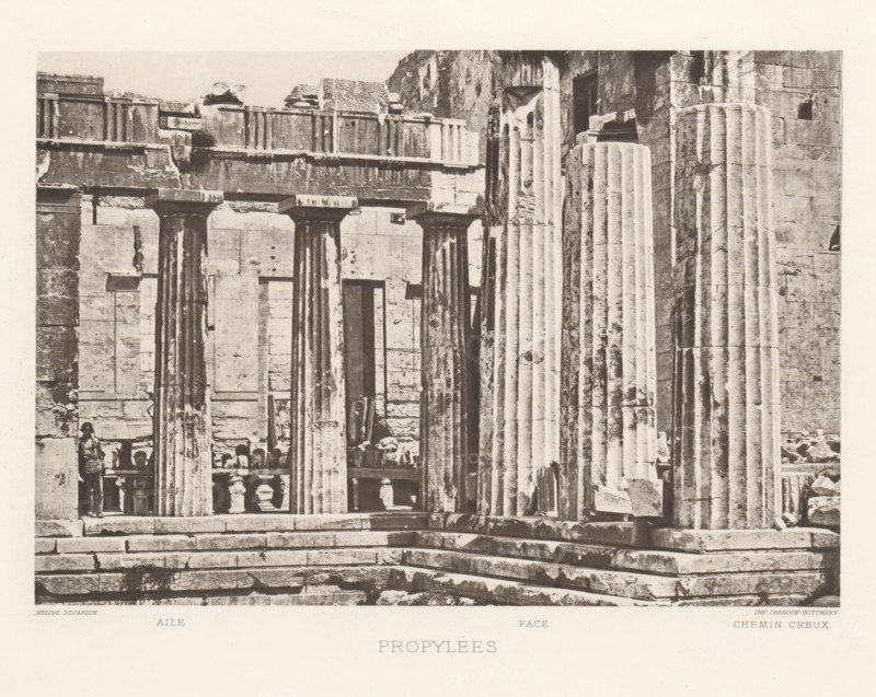 Propylées, Propylées of the Acropolis of Athens, page, photomechanical print, inscription: 'Aile Face Chemin creux', Athens, cliché maker: Dujardin, printer (person): Chardon-Wittmann, c. 1880 - in or before 1890, photographer, printer (person), Athene, Periféreia Protevoúsis, paper, photoaquatint, height 145 mm x width 215 mm, ruin of a building, architecture, column, pillar, gate, entrance, staircase, Acropolis, Propylaea (Athens), A sepia-toned photograph showing a stone entrance with tall fluted columns and a stepped platform in front, weathered block walls and lintels above the columns, a row of balustrades and sculpted fragments visible behind the columns, and a person standing near one of the columns; the stone surfaces display chips, stains, and textured aging.