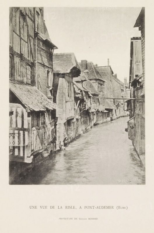 View of the Risle at Pont-Audemer, Une vue de la Risle, a Pont-Audemer (Eure), page, photomechanical print, number: 'Pl. VI.', photographer: G. Robard, Pont-Audemer, c. 1885 - in or before 1895, paper, collotype, photoprint, height 186 mm x width 144 mm, river, façade (of house or building), Risle, A narrow waterway runs between rows of closely packed timber-framed buildings with steep, uneven roofs and projecting upper floors, the façades showing visible beams, plaster, and brickwork, many windows of varying sizes and some overhanging eaves; the surface of the water appears rippled and reflective, and a simple wooden footbridge spans the channel between the buildings further along, while chimneys and rooflines create a jagged skyline against a pale sky.
