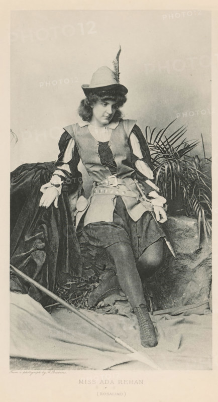 Miss Ada Rehan (Rosalind), page, photomechanical print, photographer: Alexander Bassano, cliché maker: anonymous, c. 1880 - in or before 1890, paper, photoaquatint, height 203 mm x width 135 mm, Person, Ada Rehan, portrait of actor, actress, A seated person wearing a feathered hat and a costume with laced bodice and puffed sleeves appears in a monochrome photograph, resting one arm on a draped dark fabric and the other on a stone ledge near a cluster of long, narrow-leaved plants; the outfit includes a wide belt with attached pouches, a short skirt over fitted stockings, and laced ankle boots, and a long staff or pole lies diagonally across the foreground while fallen leaves and textured fabric cover the ground.