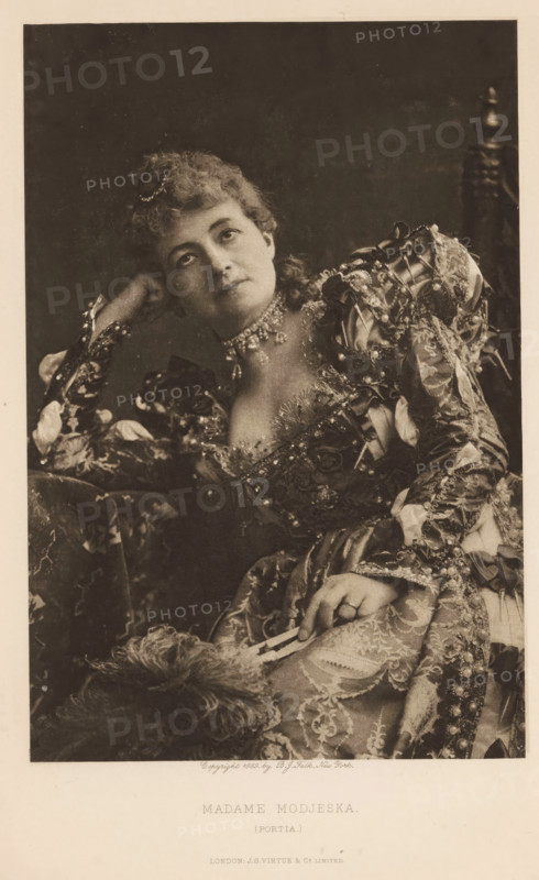 Madame Modjeska (Portia), page, photomechanical print, photographer: Benjamin Joseph Falk, New York (city), cliché maker: anonymous, publisher: Virtue & Co., London, c. 1880 - in or before 1890, North America, paper, photoaquatint, height 206 mm x width 137 mm, Person, Helena Modjeska, portrait of actor, actress, A seated woman wearing an ornate, patterned dress with puffed, decorated sleeves and lace details leans her head on one hand and looks slightly upward; she has curled hair adorned with a small hair ornament and wears a multi-strand necklace with dangling elements; her other hand rests on her lap holding a folded fan, and a feathered accessory lies nearby; the background is dark and plain and a carved wooden chair back is visible behind her.