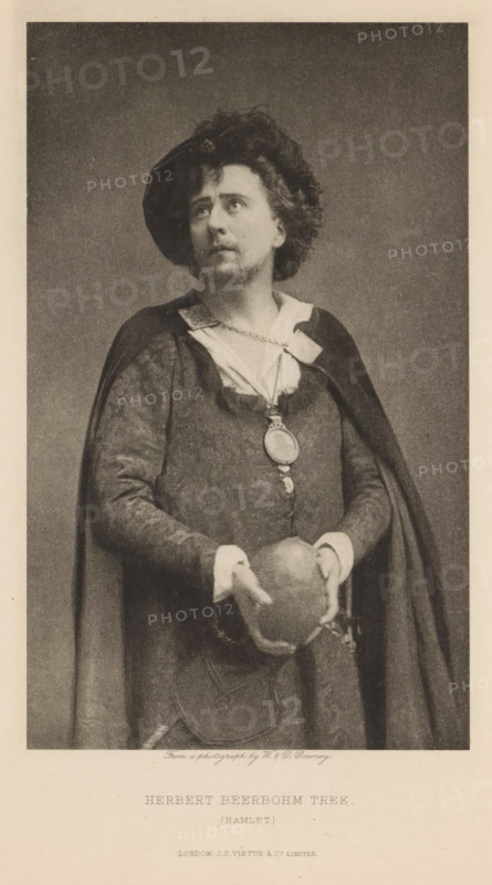 Herbert Beerbohm Tree (Hamlet), page, photomechanical print, photographer: Downey, William & Daniel, cliché maker: anonymous, publisher: Virtue & Co., London, c. 1880 - in or before 1890, paper, photoaquatint, height 198 mm x width 121 mm, Person, portrait of actor, actress, A sepia-toned portrait of a man standing and facing slightly to his left while looking upward, wearing a dark cape over a textured tunic with a light collar and cuffs, a pendant hanging on a chain at his chest, and a soft hat tilted on his curly hair; he holds a round object with both hands at waist level against a plain background.