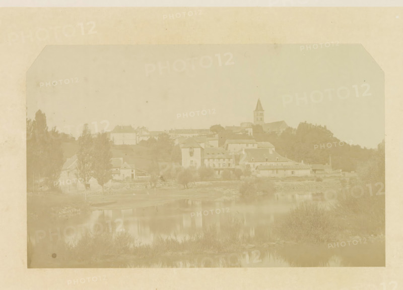Norman Keep or Caesar's Tower (before 1133), from the south-east, View of the ruins of Caesar's Tower of Kenilworth Castle, photograph, page, Kenilworth Castle, c. 1862 - in or before 1872, photographer, Kenilworth, paper, albumen print, height 152 mm x width 202 mm, ruin of a dwelling, house, castle, tower, clock tower, monumental adornment, A faded, sepia-toned panoramic view shows a riverside scene with reflections in calm water, foreground vegetation along the riverbank, clusters of trees, and a series of buildings of varying sizes and roof shapes lining the water’s edge, including a taller structure with a pointed steeple rising above the other rooftops; the image is bordered by a lighter margin and has a soft, aged appearance with faint spots and uneven tonality.