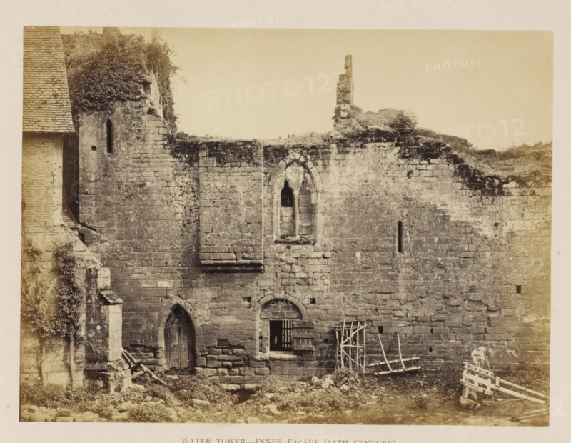 Inner wall of the ruin of a water tower of Kenilworth Castle, Water Tower - Inner façade (13th Century), photograph, page, Kenilworth Castle, c. 1862 - in or before 1872, photographer, Kenilworth, paper, albumen print, height 152 mm x width 204 mm, ruin of a dwelling, house, castle, water tower, A weathered stone wall with irregular rectangular blocks, showing areas of darker discoloration and patches of vegetation growing on and over the upper edge, contains a narrow arched window high up and a larger pointed arched opening below with a wooden door set into it, a square barred window with a wooden shutter sits beside the lower arch, and leaning wooden ladders and racks rest against the wall near the ground amid scattered stones and rubble.