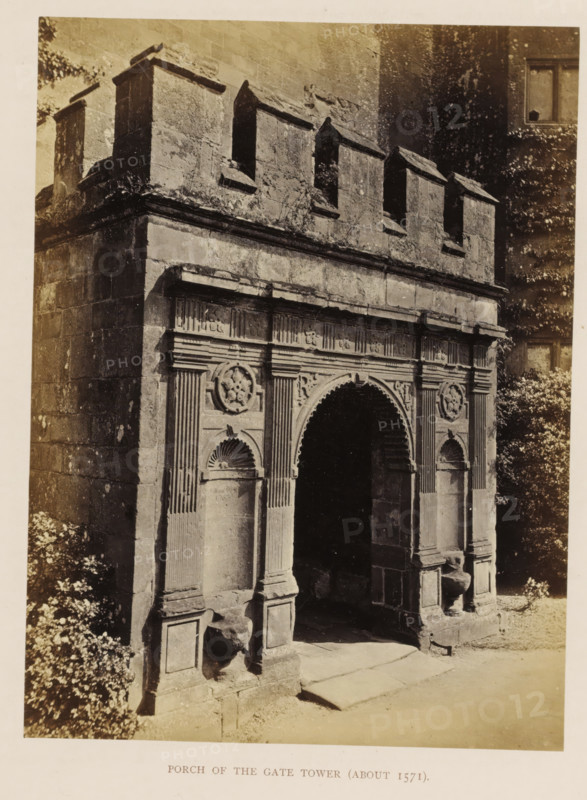 Gate of a tower of Kenilworth Castle, Porch of the Gate Tower (about 1571), photograph, page, Kenilworth Castle, c. 1862 - in or before 1872, photographer, Kenilworth, paper, albumen print, height 203 mm x width 152 mm, ruin of a dwelling, house, castle, monumental door, porch, 'aediculum', A sepia photograph of a stone gate porch showing an arched entrance flanked by decorated pilasters and recessed niches, above which runs a horizontal entablature with carved panels and circular floral medallions; the upper section features a crenellated parapet with rectangular embrasures, and the stone surfaces show weathering and patches of lichen; foliage is visible at the sides of the structure and a shadowed interior beyond the arch contrasts with the sunlit stone step and ground in front.