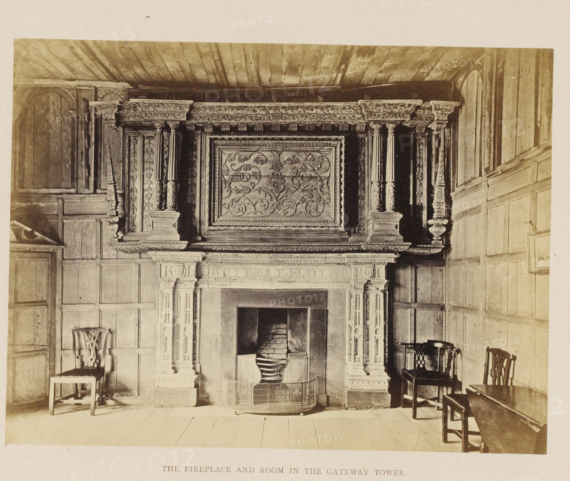 Fireplace in Kenilworth Castle, The Fireplace and Room in the Gateway Tower, photograph, page, Kenilworth Castle, c. 1862 - in or before 1872, photographer, Kenilworth, paper, albumen print, height 152 mm x width 202 mm, open hearth, fire-place, ruin of a dwelling, house, castle, A large carved stone fireplace with an ornate overmantel featuring floral and scrollwork panels is centered in a paneled room, flanked by decorative pilasters and carved columns supporting a heavily carved frieze. The fireplace opening shows stacked bricks and a metal fireguard curved around the hearth. Wooden wall paneling covers the surrounding walls and a plank ceiling runs overhead. Simple wooden chairs with carved backs and a small wooden table are placed near the fireplace on a wide wooden plank floor.