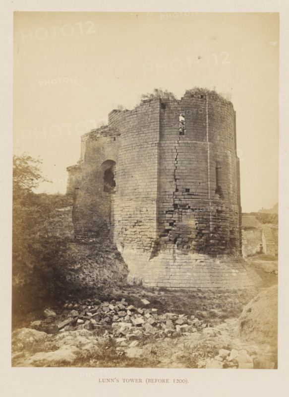 Ruin of Lunn's Tower of Kenilworth Castle, Lunn's Tower (before 1200), photograph, page, Kenilworth Castle, c. 1862 - in or before 1872, photographer, Kenilworth, paper, albumen print, height 204 mm x width 152 mm, ruin of a dwelling, house, castle, tower, clock tower, monumental adornment, A tall circular stone tower with a battered base and visible horizontal courses shows areas of missing and crumbling masonry and a narrow vertical slit opening with vegetation poking through; adjacent to it is a partially ruined wall with an arched gap and jagged top, and rubble and scattered stones lie on grassy earth in the foreground while low vegetation and a tree appear to the left.