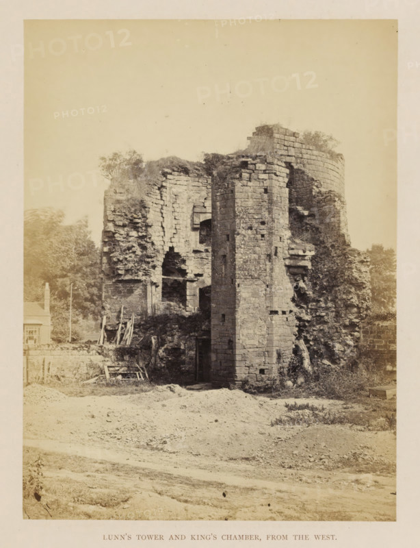 Lunn's Tower and King's Chamber, from the west, View of the ruins of Lunn's Tower and the king's departure from Kenilworth Castle, photograph, page, Kenilworth Castle, c. 1862 - in or before 1872, photographer, Kenilworth, paper, albumen print, height 204 mm x width 152 mm, ruin of a dwelling, house, castle, tower, clock tower, monumental adornment, A sepia-toned photograph of a crumbling stone tower and adjoining ruined walls set on rough ground, showing weathered masonry blocks, arched openings, and jagged collapsed sections. Vegetation grows atop parts of the structure and around its base, with scattered wooden planks and construction remnants leaning against a low wall nearby. Trees and a small building appear in the background to one side, and the foreground shows uneven dirt and sparse grassy patches leading up to the ruin.