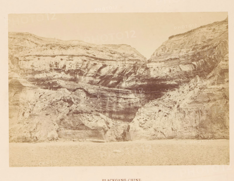 Blackgang Chine, photograph, page, photographer: Frank Mason Good, Isle of Wight, c. 1859 - in or before 1869, paper, albumen print, height 100 mm x width 144 mm, rocky coast, coastal cliff or crag, A sepia-toned photograph of a steep rocky cliff face with layered horizontal strata and eroded textures, showing dark shadowed recesses and lighter weathered surfaces; a narrow stepped path carved into the rock leads up from the lower central area into a small dark opening; the foreground is a flat sandy or pebbled beach area, and the upper portion of the image shows a pale sky above the cliff rim.