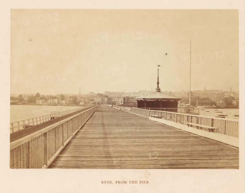 Ryde, from the pier, photograph, page, photographer: William Russell Sedgfield, c. 1859 - in or before 1869, Isle of Wight, paper, albumen print, height 101 mm x width 144 mm, city-view in general, 'veduta', pier, quay, wharf, A wide wooden pier extends toward a distant shoreline, with wooden planks forming the walking surface and railings running along both sides; a pavilion with a pointed central finial and a column rises on the right side of the pier, and a tall thin mast stands beyond it; the shoreline shows rows of buildings and trees with various rooflines and a few church spires faintly visible against a pale sky; calm water borders the left side of the pier and small boats appear near the right shoreline; the overall image has a sepia tone and an aged, slightly faded appearance.