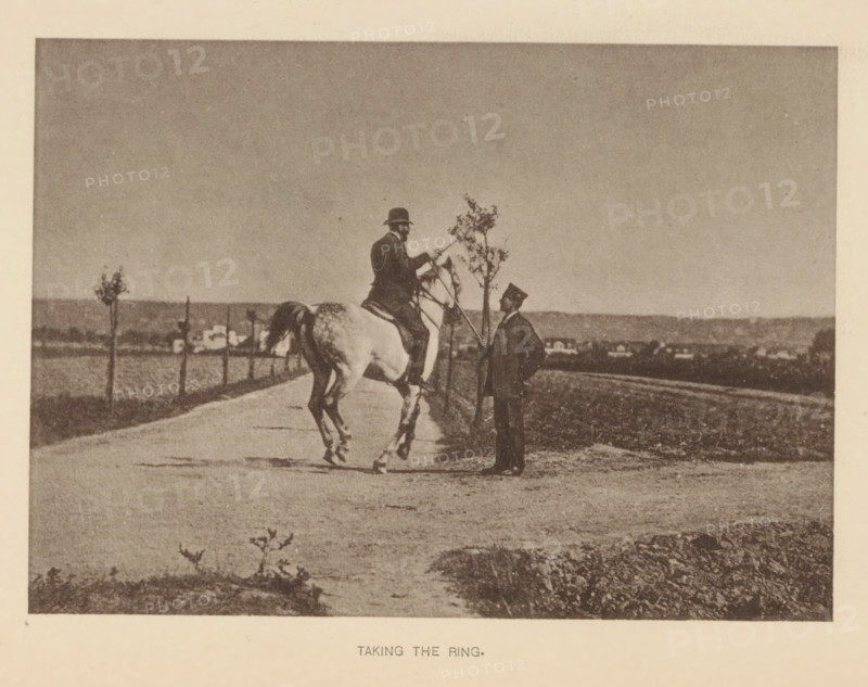 Man riding a horse and presumably participating in ring jousting, Taking the ring, page, photomechanical print, cliché maker: Römmler & Jonas, Dresden, c. 1879 - in or before 1889, photographer, paper, collotype, photogravure, height 100 mm x width 140 mm, horse, riding a horse, ass, or mule, rider, horseman, head-gear: bowler hat, A sepia-toned photograph showing a person seated on a light-colored horse on a road, the rider wearing a hat and leaning slightly forward while holding the reins, facing another standing person who is dressed in a dark suit and hat and holding a long pole or stick near a small tree, a line of small trees and a distant low horizon with scattered buildings visible in the background, with the foreground showing a textured dirt or gravel surface and some low vegetation.