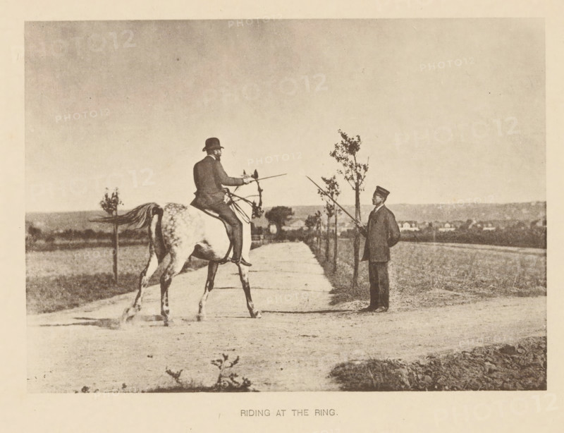 Man riding a horse and presumably going to joust, Riding at the ring, page, photomechanical print, cliché maker: Römmler & Jonas, Dresden, c. 1879 - in or before 1889, photographer, paper, collotype, photogravure, height 97 mm x width 136 mm, horse, riding a horse, ass, or mule, rider, horseman, head-gear: bowler hat, A sepia-toned photograph shows a man seated on a horse facing a second man standing on a dirt path lined with small trees; the rider wears a dark coat and a hat and holds a long, thin pole toward the standing man, who is dressed in a dark suit and cap and also holds a pole; the horse is light-colored with visible markings and a raised tail, and the background features open fields and distant low buildings under a wide, empty sky.