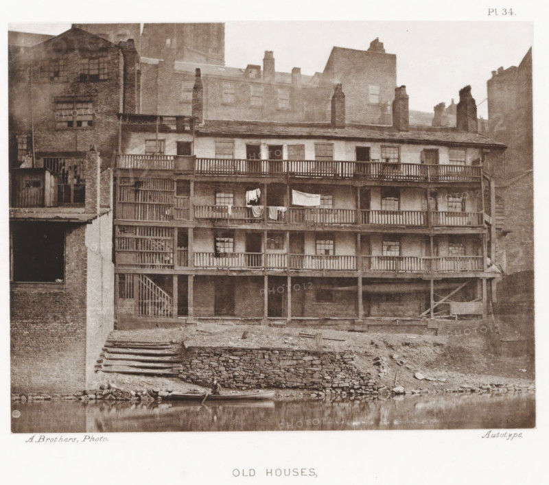 View of a house in Manchester with an open gallery, Old houses, parsonage, page, photomechanical print, number: 'Pl. 34.', photographer: Brothers, Alfred, Manchester, cliché maker: anonymous, c. 1868 - in or before 1878, printer (person), paper, collotype, photoprint, height 140 mm x width 179 mm, Print after page 30., housing, A multi-story brick building with wooden balconies running across several levels, each balcony supported by vertical posts and accessed by an exterior wooden staircase, clothes hanging over one railing, multiple small paned windows set into the facade, chimney stacks rising from the roofline, adjoining taller brick buildings visible behind and beside it, a stone terraced embankment leading down to a calm body of water in the foreground with a small boat pulled up at the edge, exposed earth and scattered debris along the shoreline.