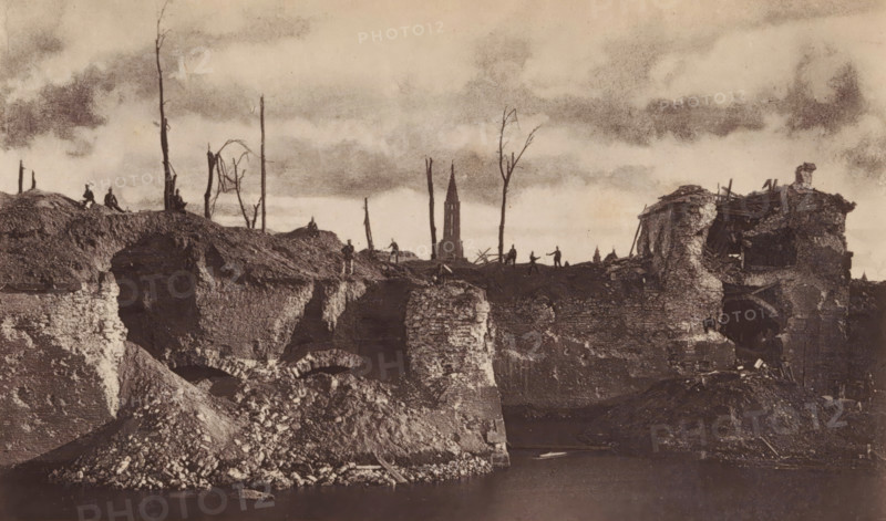 View of a breach in a bastion in Strasbourg as a result of the siege by the German army, Face gauche de la brèche du bastion 12 et porte de pierres, page, photomechanical print, photographer: Charles David Winter, Strasbourg (France), cliché maker: anonymous, printer (person): Goupil & Cie., 1870, printer (person), paper, Woodburytype, height 101 mm x width 170 mm, Print on frontispiece., landscape with ruins, assault, siege, consequences of war, A wide, sepia-toned scene shows a ruined landscape with crumbling stone walls and mounds of rubble descending to a dark body of water in the foreground, jagged exposed brick and arch shapes visible in the ruins. On the higher ground above the ruins are silhouettes of bare, leafless tree trunks and sparse branches rising against a cloudy sky. Small human figures stand and sit along the ridge, their forms dark and indistinct. On the right a partially collapsed building with broken beams and torn masonry leans over the edge of the ruin, with scat...