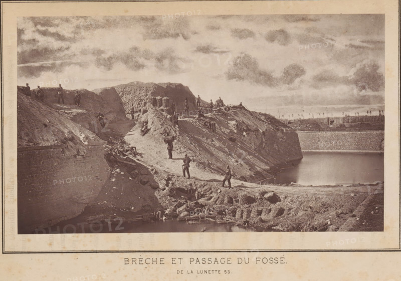 View of a breach and a passage through a fortress in Strasbourg, Brèche et passage du fossé de la lunette 53, page, photomechanical print, photographer: Charles David Winter, Strasbourg (France), cliché maker: anonymous, printer (person): Goupil & Cie., 1870, printer (person), paper, Woodburytype, Woodburytypepie, height 101 mm x width 171 mm, landscape with ruins, assault, siege, consequences of war, A sepia-toned image shows a rugged earthen embankment descending to a water-filled trench with a high stone retaining wall along its far edge; the embankment is uneven and eroded with exposed soil and scattered debris, and woven baskets or gabions are stacked near the lower edge by the water. Several uniformed figures stand, sit, and walk along the slope and crest, some carrying packs or equipment and one appearing to lean on a long object, while a few others are positioned near the top beside earthen fortifications and stacked cylindrical shapes. In the background a line of wooden stakes...
