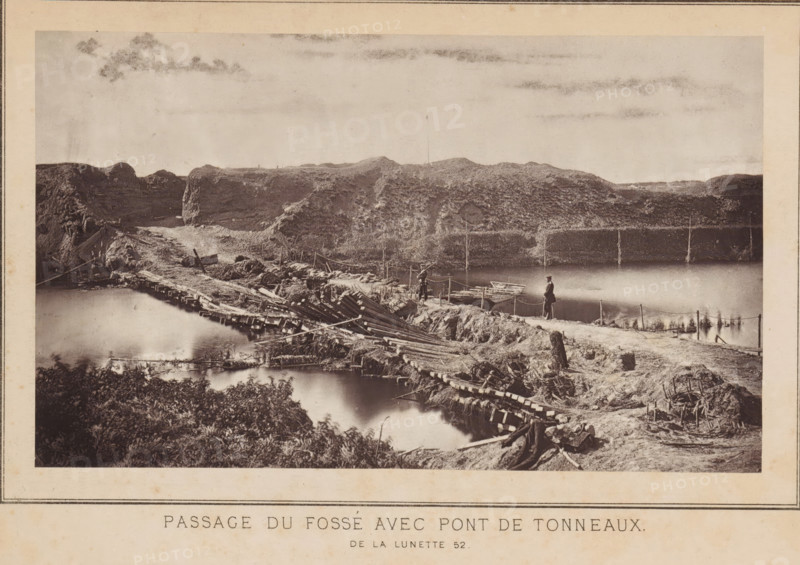 Passage du fossé avec pont de tonneaux de la lunette 52, View of a passage through a fortress and a pontoon bridge in Strasbourg, page, photomechanical print, photographer: Charles David Winter, Strasbourg (France), cliché maker: anonymous, printer (person): Goupil & Cie., 1870, printer (person), paper, Woodburytype, Woodburytypie, height 101 mm x width 171 mm, assault, siege, pontoon bridge, consequences of war, A sepia-toned photograph showing a rough wooden bridge and pathway crossing a body of water with reflections, flanked by earthworks and uneven terrain; the foreground includes dense foliage and scattered wooden debris, while the middle ground features men standing on the path near small boats tied at the water’s edge and ropes or posts lining the route; the background shows rocky, grassy embankments rising to a ridge under a sky with light cloud streaks.