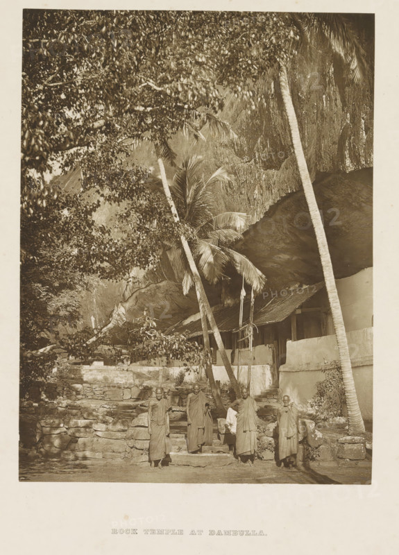 Exterior of the Dambulla Cave Temple, Sri Lanka, page, photomechanical print, Four monks stand in the foreground., title: 'ROCK TEMPLE AT DAMBULLA.', photographer: Henry William Cave, Dambulla, cliché maker: anonymous, 1896, South Asia, paper, Woodburygravure, height 184 mm x width 137 mm, Print on second endpaper., temple, shrine, Hinduism, Buddhism, Jainism, rock-formations, temple servants, Golden temple of Dambulla, A tall rocky overhang fills the upper area with rough textured stone and hanging foliage, several slender palm trunks rising from the ground and leaning against the rock, a low stone wall and stepped stone platform in front of a simple building with a sloped thatched roof and plain plaster walls, a group of people standing on the steps and in front of the wall wearing long draped garments, dense tree branches and leaves framing the left side of the scene.