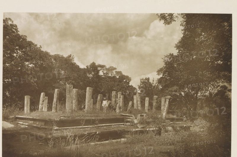 Ruins of a pavillion at Anuradhapura, Sri Lanka, page, photomechanical print, title: 'REMAINS OF PAVILLION (THIRD CENTURY).', photographer: Henry William Cave, Anuradhapura, cliché maker: anonymous, 1896, South Asia, paper, Woodburygravure, height 124 mm x width 193 mm, Print for page 79., ruin of a building, architecture, column, pillar, A sepia-toned outdoor scene shows a grassy area with a series of upright stone pillars of varying heights arranged in a loose formation, surrounded by low stone foundations and scattered rectangular stone blocks. A solitary person stands among the pillars wearing light clothing and a head covering. Dense trees frame the background on both sides, and a cloudy sky fills the upper portion of the image. The ground in the foreground has grasses and low vegetation interspersed with the stone structures.
