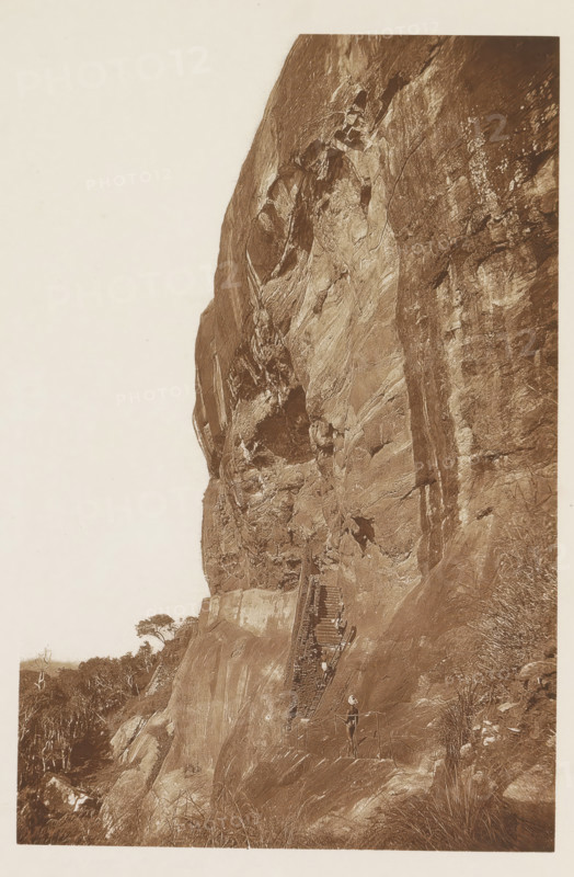 View of the gallery at the Lion Rock, Sigiriya, Sri Lanka, page, photomechanical print, title: 'REMAINS OF THE GALLERIES OF SIGIRI.', photographer: Henry William Cave, cliché maker: anonymous, 1896, South Asia, paper, Woodburygravure, height 191 mm x width 125 mm, Print for page 95., ruin of a dwelling, house, castle, rock-formations, A steep rock face fills most of the image, showing weathered stone surfaces with visible cracks, ledges, and textured striations. A narrow wooden staircase with railings ascends along the rock, anchored to the stone, and several small figures stand on and near the stairs. Vegetation including low grasses and scattered trees appears at the base and along the left edge of the scene. The photograph is sepia-toned and framed with a pale margin.