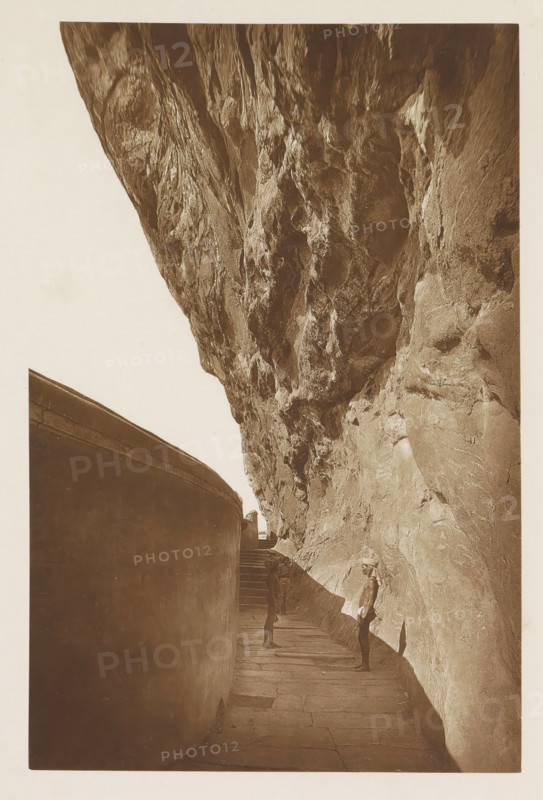 In the gallery of the Lion Rock at Sigiriya, Sri Lanka, page, photomechanical print, title: 'IN THE GALLERY OF SIGIRI.', photographer: Henry William Cave, Sigiriya, cliché maker: anonymous, 1896, South Asia, paper, Woodburygravure, height 191 mm x width 126 mm, Print for page 97., rock formations, gallery (spacious corridor), A narrow stone walkway runs between a tall, curved wall on the left and a steep, textured rock face on the right, the rock towering overhead with rough, irregular surfaces and deep crevices; two men stand along the path, one facing the other partway down the walkway and another closer to the foreground wearing a short garment and head covering, their shadows cast on the rock, while a short flight of steps leads up toward a small opening where the curved wall meets the rock.