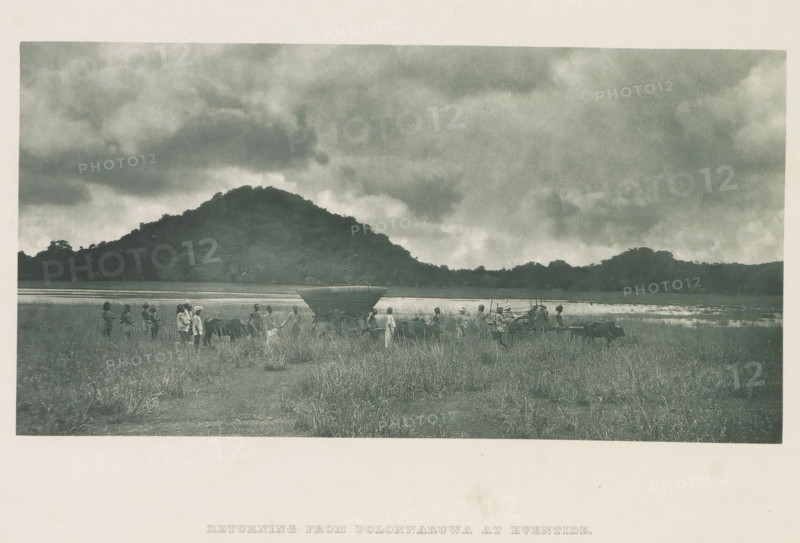 Tour group with bullock carts in Minneriya, Sri Lanka, page, photomechanical print, title: 'RETURNING FROM POLONNARUWA AT EVENTIDE.', photographer: Henry William Cave, Minneriya, cliché maker: anonymous, 1896, South Asia, paper, Woodburygravure, height 101 mm x width 196 mm, Print for page 103., 'en route', traveler under way, two-wheeled vehicle drawn by one animal, ox, A wide landscape photograph showing a grassy field in the foreground with a group of people standing and walking among two horse-drawn carts; one cart has a covered top and large wooden wheels while the other is more open and shows harnessed animals at the shafts; the people wear simple clothing and head coverings and some appear to be guiding the animals. Behind them is a strip of water reflecting the sky and a dark, tree-covered hill rising across the middle of the image. Above the hill a broad sky filled with heavy, textured clouds occupies the upper portion of the scene.
