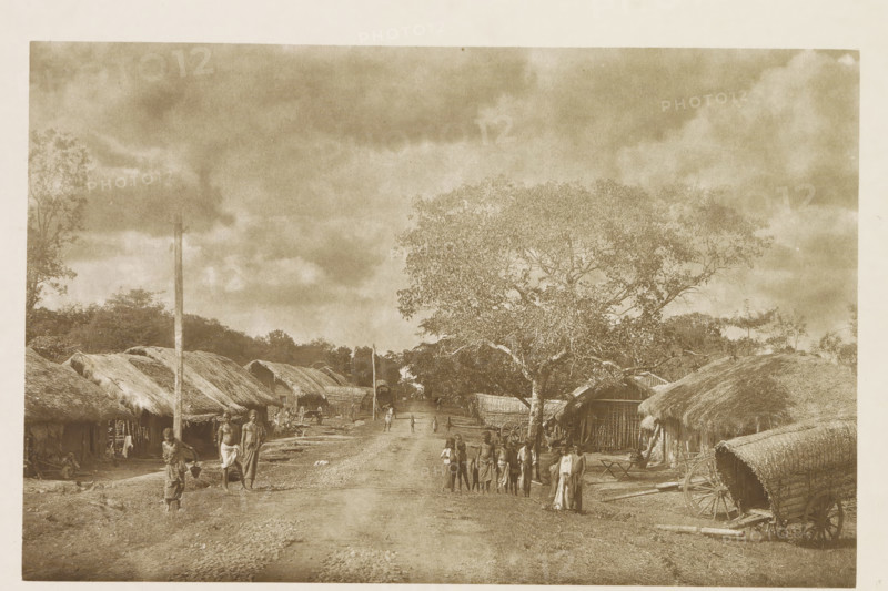 Path through Kekirawa, Sri Lanka, page, photomechanical print, title: 'KEKIRAWA.', photographer: Henry William Cave, Kekirawa, cliché maker: anonymous, 1896, South Asia, paper, Woodburygravure, height 124 mm x width 192 mm, Print for page 21., public road in village, village, A sepia-toned scene shows a dirt road flanked by thatched huts with woven walls and low roofs, a large leafy tree on the right side of the road, and a wooden cart with a curved thatched cover parked near the right foreground; scattered people in traditional clothing stand and walk along the road and near the huts, including small groups and individuals carrying items, while a tall pole rises near the left side and distant trees and a cloudy sky fill the background.