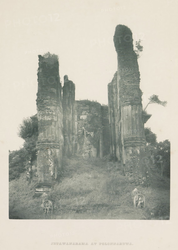 Ruins of the Lankatilaka Vihara at Polonnaruwa, Sri Lanka, page, photomechanical print, title: 'JETAWANARAMA AT POLONNARUWA', photographer: Henry William Cave, Polonnaruwa, cliché maker: anonymous, 1896, South Asia, paper, Woodburygravure, height 196 mm x width 146 mm, Print for page 109., temple, shrine, Hinduism, Buddhism, Jainism, ruin of a building, architecture, A tall stone ruin rises from a grassy slope, consisting of several weathered vertical pillars and walls with layered textures and eroded surfaces, some topped with patches of vegetation and small trees; two sculpted stone figures sit near the base of the slope facing forward, and bushes and foliage grow around the lower parts of the structure, all shown against a pale, empty sky.