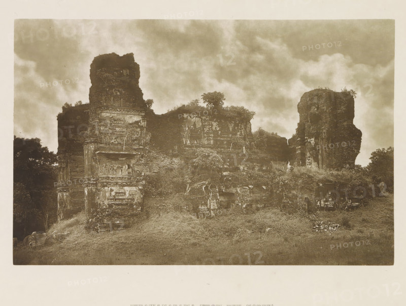 Ruins of the Lankatilaka Vihara at Polonnaruwa, Sri Lanka, page, photomechanical print, title: 'JETAWANARAMA (FROM THE NORTH).', photographer: Henry William Cave, Polonnaruwa, cliché maker: anonymous, 1896, South Asia, paper, Woodburygravure, height 126 mm x width 195 mm, Print for page 111., temple, shrine, Hinduism, Buddhism, Jainism, ruin of a building, architecture, A sepia-toned photograph showing a large ruined stone structure with weathered walls and crumbling towers rising against a cloudy sky, vegetation and small trees growing on and around the masonry, carved stone details and niches visible on the facade, grassy ground in front with scattered stones and low plants, and dense foliage at the edges of the composition.