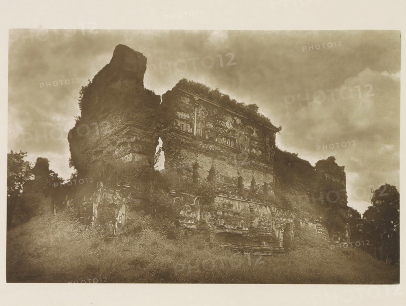 Ruins of the Lankatilaka Vihara at Polonnaruwa, Sri Lanka, page, photomechanical print, title: 'JETAWANARAMA (FROM THE SOUTH-WEST).', photographer: Henry William Cave, Polonnaruwa, cliché maker: anonymous, 1896, South Asia, paper, Woodburygravure, height 126 mm x width 195 mm, Print for page 111., temple, shrine, Hinduism, Buddhism, Jainism, ruin of a building, architecture, A sepia-toned photograph showing a large, crumbling stone structure with weathered, layered masonry and decorative reliefs partially obscured by vegetation; the ruin rises against a cloudy sky with plants and vines growing on and around its upper edges, and the foreground shows uneven ground with grass and scattered stones.