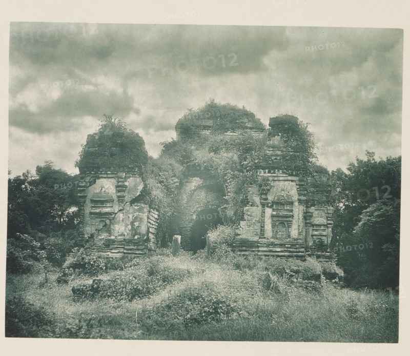 Entrance to the Thuparama temple at Polonnaruwa, Sri Lanka, page, photomechanical print, title: 'ENTRANCE TO THE THUPARAMA AT POLONNARUWA.', photographer: Henry William Cave, Polonnaruwa, cliché maker: anonymous, 1896, South Asia, paper, Woodburygravure, height 148 mm x width 186 mm, Print for page 113., temple, shrine, Hinduism, Buddhism, Jainism, ruin of a building, architecture, An overgrown stone structure with weathered, carved facades stands amidst tall grass and dense vegetation, its central archway partly obscured by plants and rubble, a terrace of steps and decorative stone elements visible on either side, with foliage and vines covering the upper sections and surrounding trees framing the ruin beneath a cloudy, textured sky.