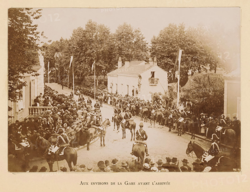 View of the Bergerac station area before the arrival of President Félix Faure, Aux environs de la Gare avant l'arrivée, photograph, page, Bergerac, 1895-06-04, photographer, paper, daylight gelatin silver print, height 118 mm x width 167 mm, Photo on frontispiece., riding a horse, ass, or mule, rider, horseman, street, triumphal entry and public reception, pageant, 'entrée solennelle', 'joyeuse entrée', A sepia-toned scene of a crowded street lined with tall leafy trees and low buildings, with many people gathered along both sides of the road watching a procession of mounted soldiers in dark uniforms and plumed helmets riding along the street, some soldiers facing forward and some seen from behind, horses wearing decorated saddles and cloths, a small light-colored house with a window box and a person standing at the doorway, tall flagpoles with long narrow banners, spectators wearing hats and coats packed closely on sidewalks and balconies, a few individuals peering from windows, and a...