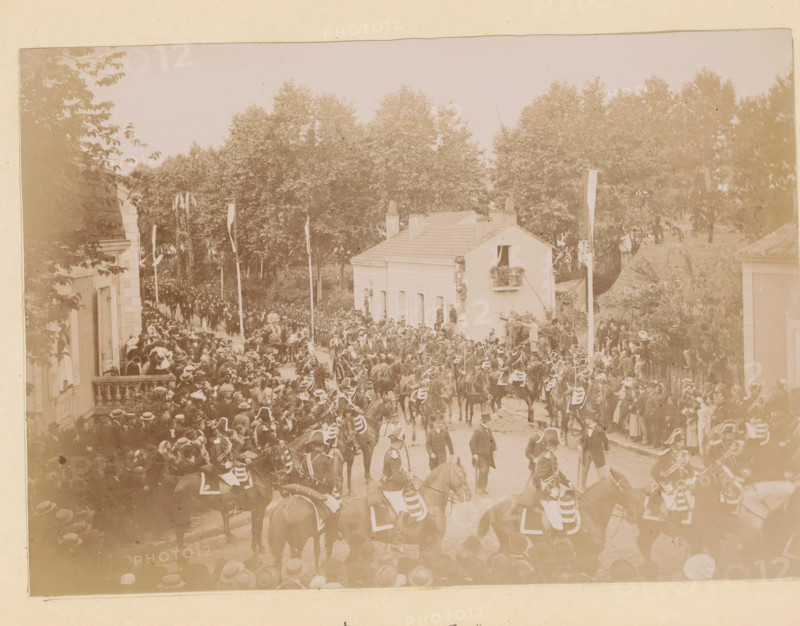 View of the arrival of President Félix Faure at Bergerac station, Départ de la gare, photograph, page, Bergerac, 1895-06-04, photographer, paper, daylight gelatin silver print, height 118 mm x width 167 mm, riding a horse, ass, or mule, rider, horseman, street, triumphal entry and public reception, pageant, 'entrée solennelle', 'joyeuse entrée', A wide street scene showing a large crowd gathered along both sides of a tree-lined avenue with tall leafy trees in the background, a small pale building with a balcony and window boxes at the center right, and several vertical banners on poles rising above the crowd; numerous horse-mounted riders in dark uniforms and plumed hats form a procession through the street, with many people standing and sitting on balconies and at street level watching, some wearing hats; to the left a stone building with a balustrade is partially visible and to the right a thatched or earth-covered embankment and another building edge can be seen, all rendered in a s...