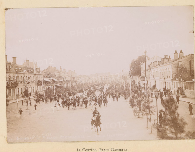 Le cortège, Place Gambetta, Bergerac- Voyage presidentiel- Place Gambetta, View of the entry of President Félix Faure on the Place Gambetta in Bergerac, photograph, page, Bergerac, 1895-06-04, photographer, paper, daylight gelatin silver print, height 118 mm x width 167 mm, riding a horse, ass, or mule, rider, horseman, square, circus, triumphal entry and public reception, pageant, 'entrée solennelle', 'joyeuse entrée', façade (of house or building), A wide street scene with rows of multi-story buildings on both sides and crowds filling the roadway and sidewalks; many people stand on balconies and at windows while a large mounted procession of riders moves down the center of the street, visible horses and riders at the front and more figures following; trees and lamp posts line the sidewalks, and decorative flags or banners hang from some buildings and poles; architectural details of the buildings include chimneys, pitched roofs, and rows of windows, and clusters of people gather near ...